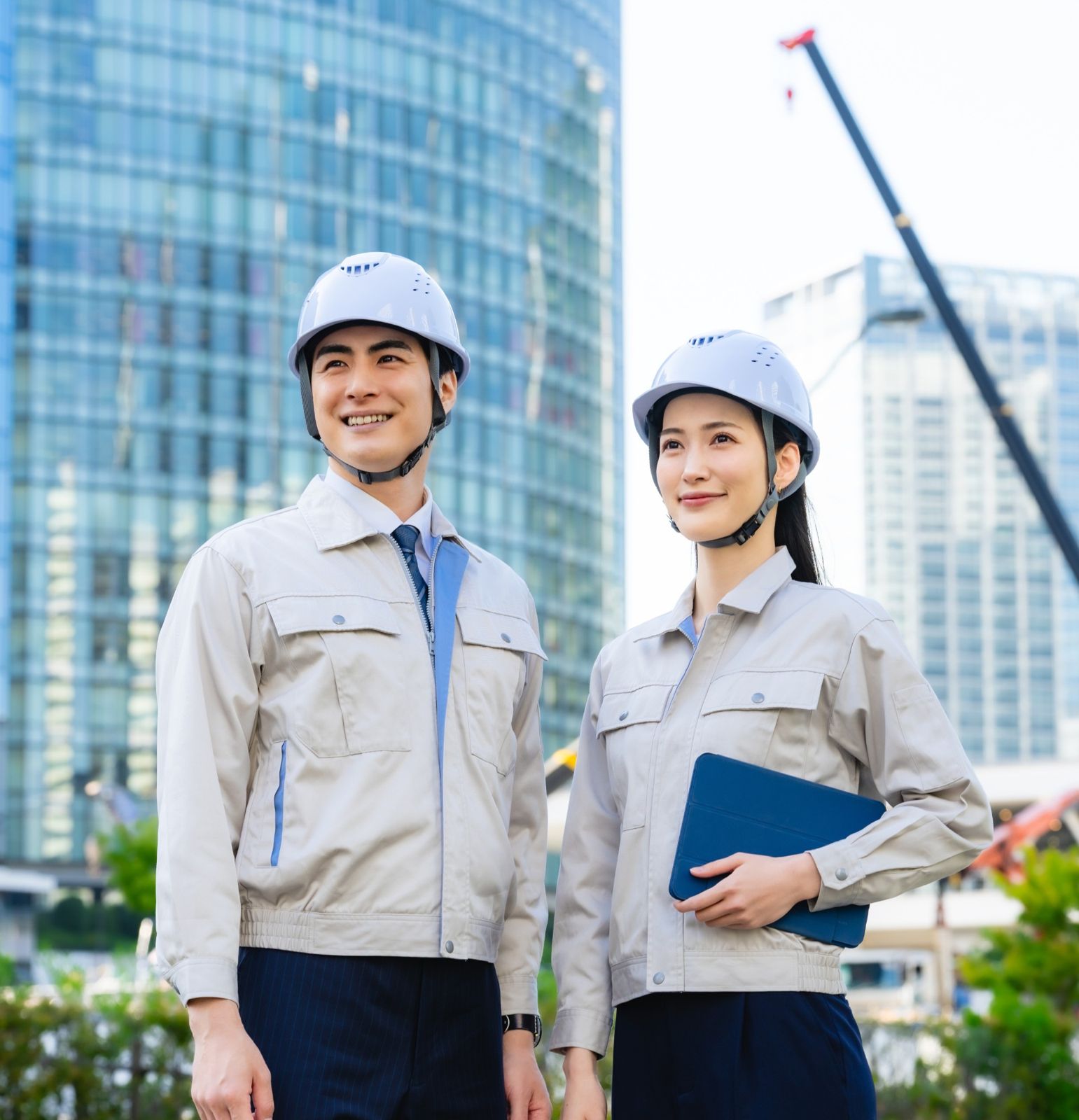 ヘルメットと作業服の男性と女性が、ビルの建設現場を背に笑顔で立っている様子